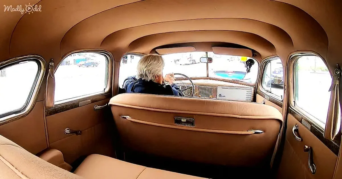 A person with white hair sits in the driver's seat of a vintage car with tan upholstery and a curved dashboard.