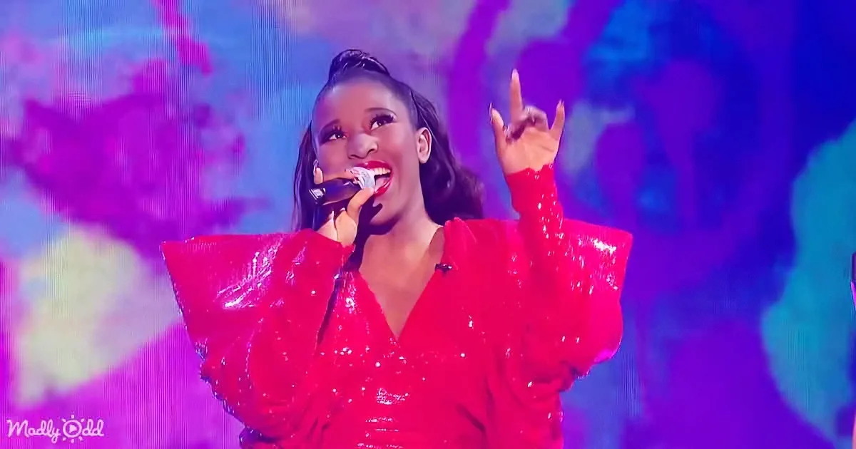 A performer in a red sequined outfit sings into a microphone, pointing upwards against a vibrant purple and blue background.