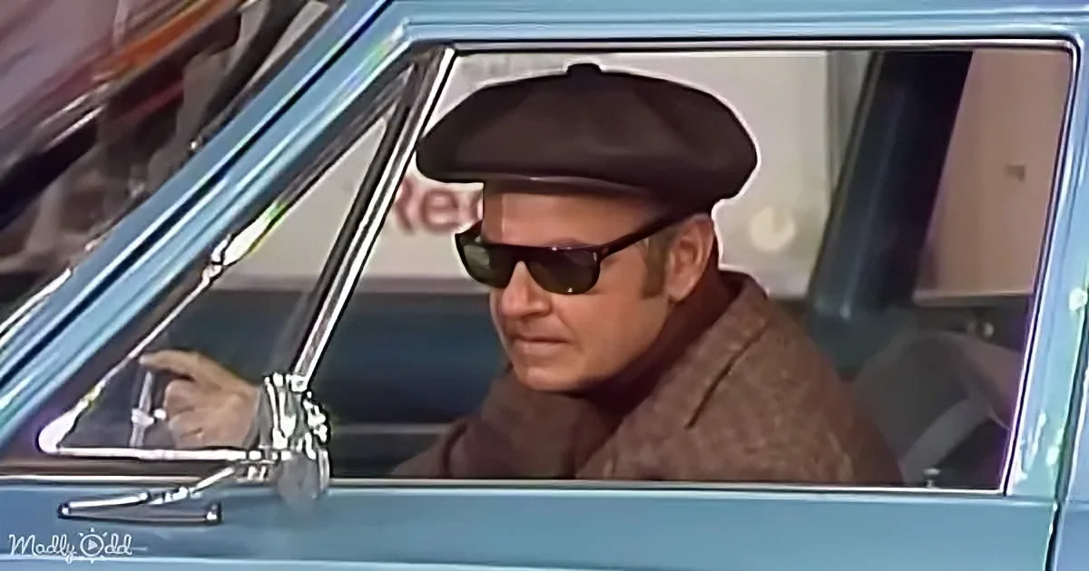 A man wearing a beret and sunglasses drives a classic car, looking serious.