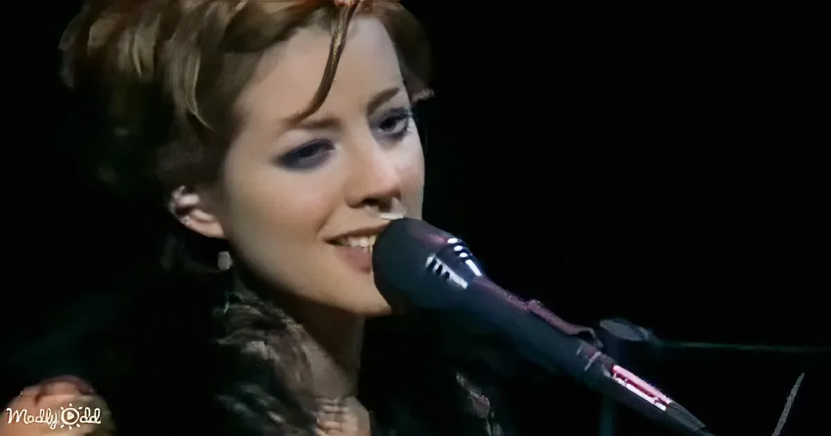 A woman with short hair sings into a microphone, wearing a dark outfit.