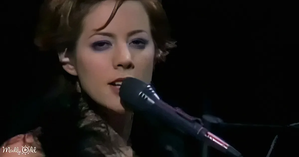 A woman with short hair sings into a microphone, wearing dark makeup and a black top.