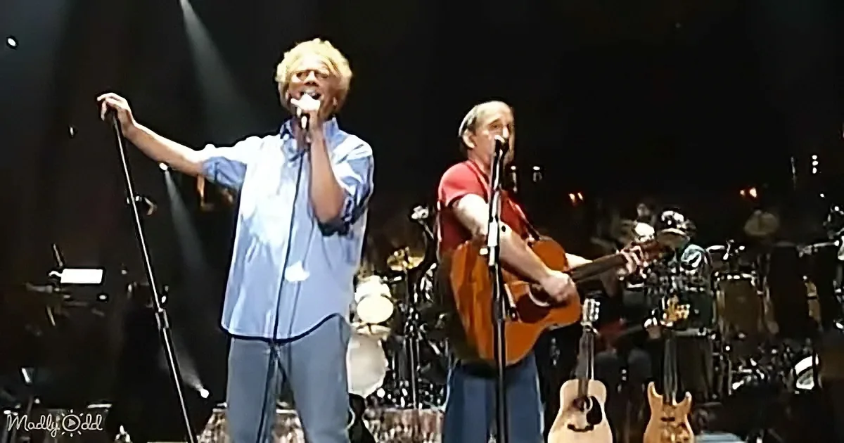 A man in a blue shirt sings into a microphone on stage, accompanied by another man playing guitar.