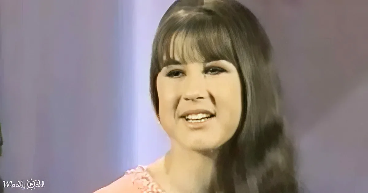 A smiling woman with shoulder-length hair and bangs, wearing a pink top, against a blurred background.