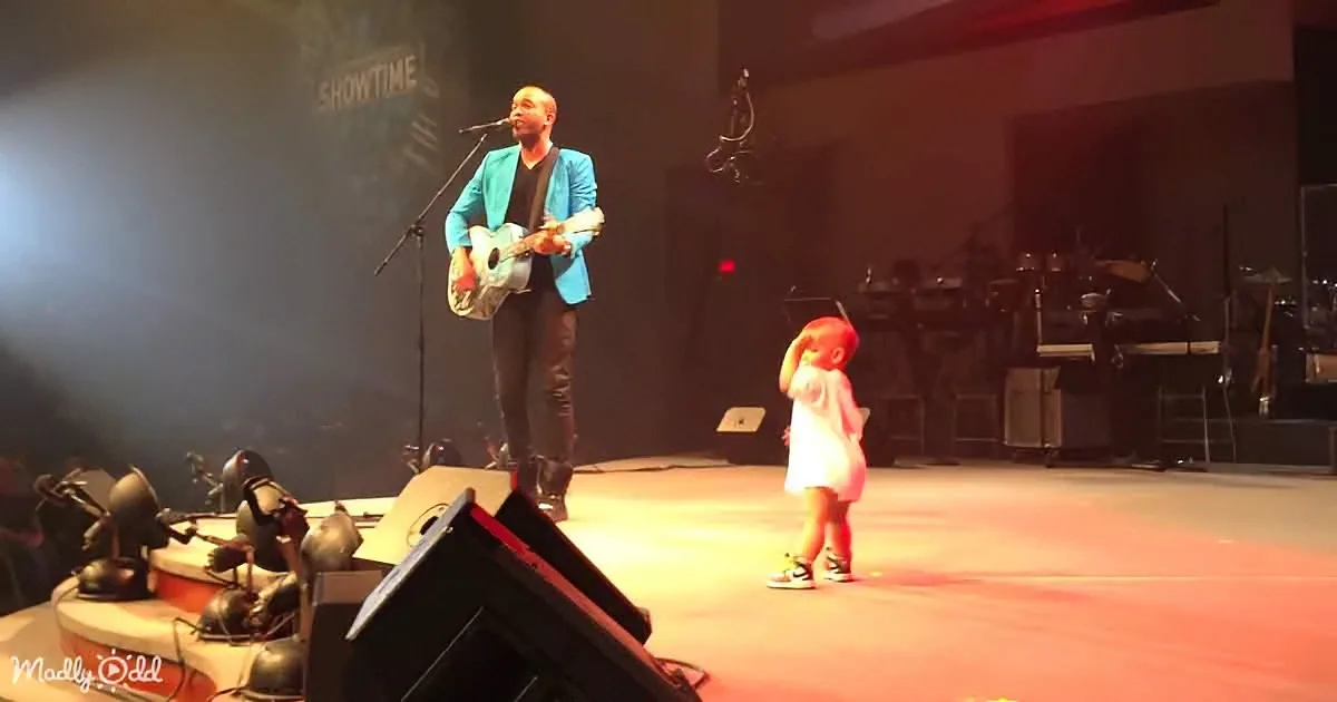 A man in a blue jacket plays guitar on stage, while a child in a pink dress stands nearby, both under stage lights.
