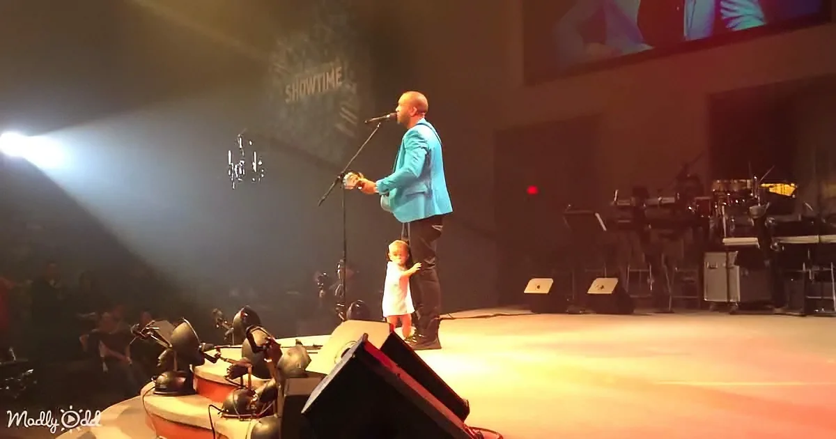 A man in a blue suit stands on stage with a child, holding a microphone, under bright stage lights.
