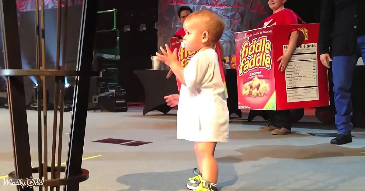 A toddler in white clothing stands on stage, holding a box labeled "Fiddle Faddle," with another child in the background.