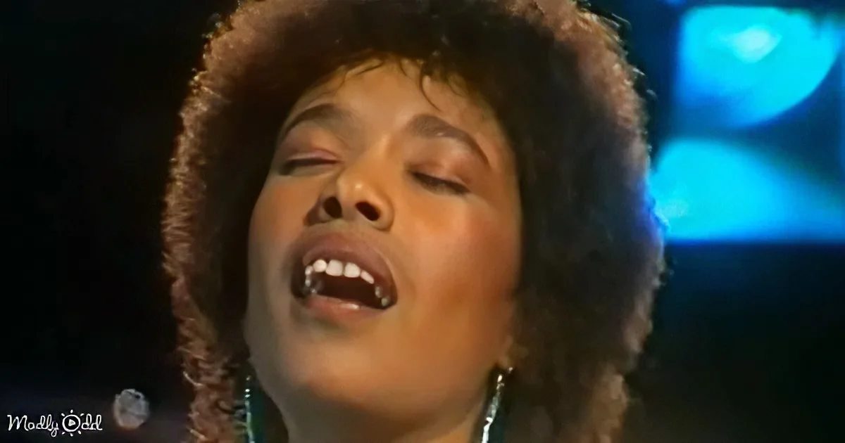 A person with curly hair and closed eyes, possibly singing, with a microphone in the background.