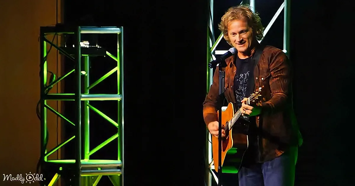 A man with curly hair plays an acoustic guitar on stage with green lighting.
