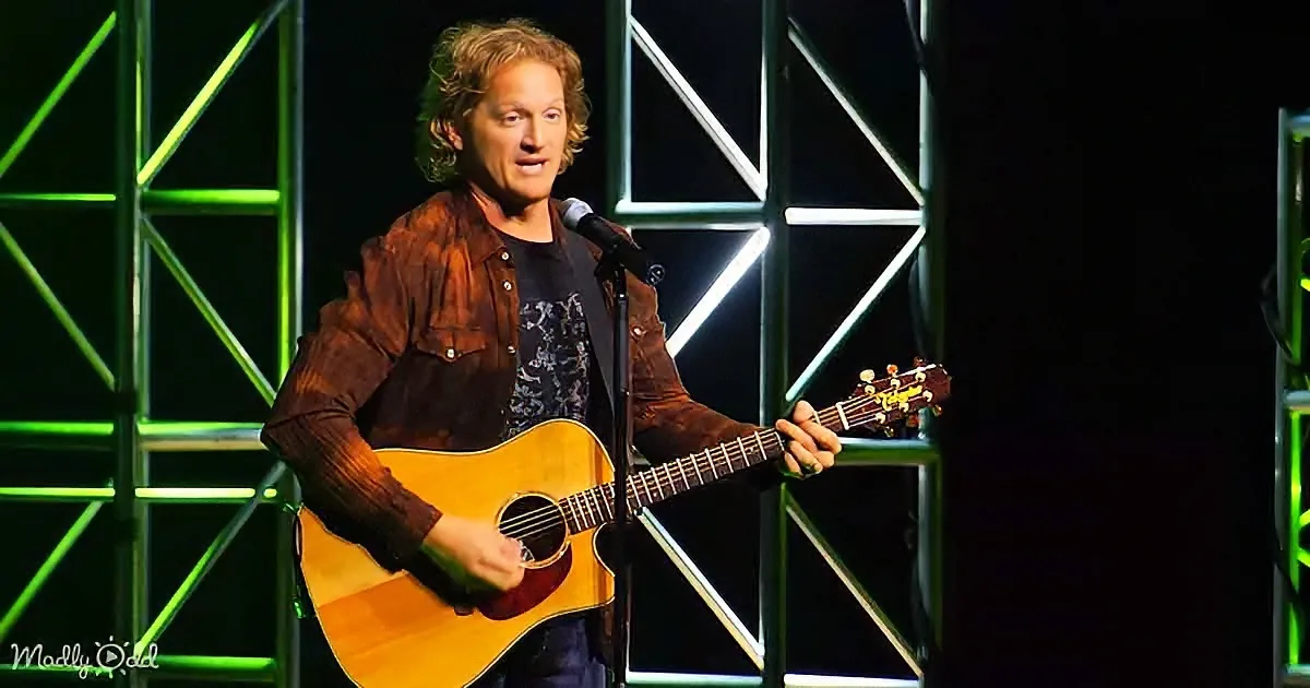 A man with curly hair plays an acoustic guitar on stage, wearing a brown shirt and black pants.