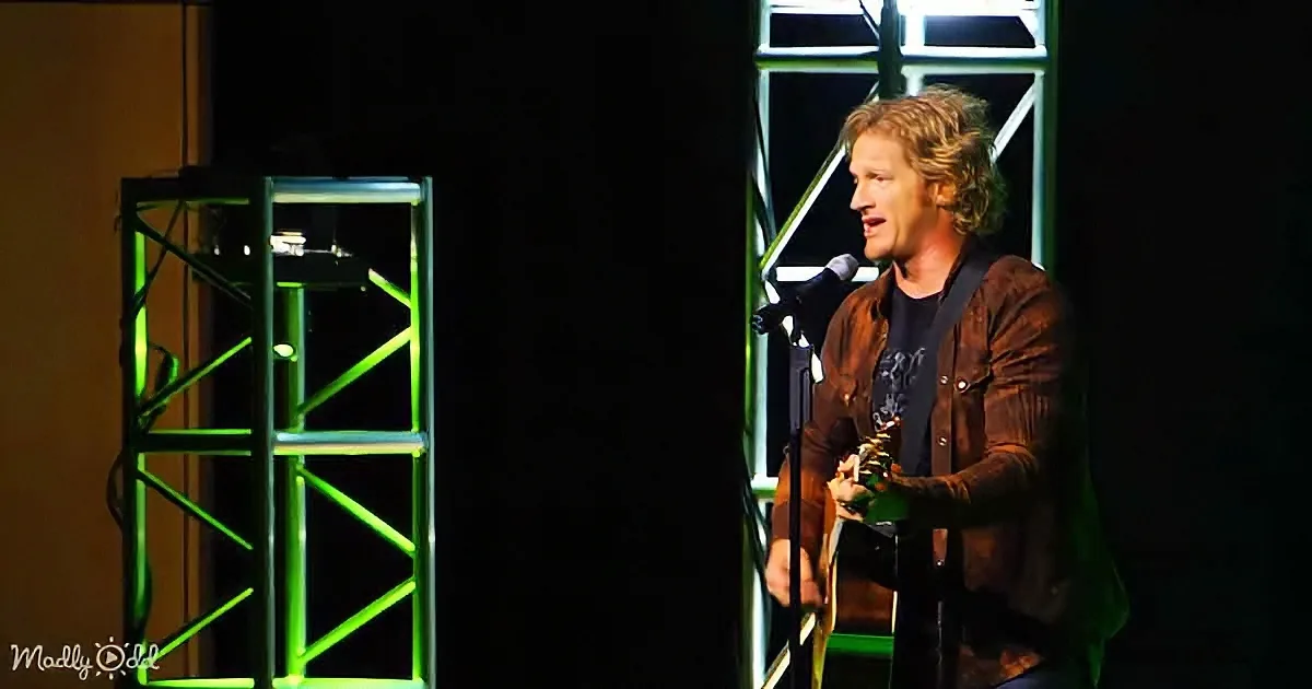 A man with curly hair, wearing a brown jacket, plays a guitar and sings into a microphone on stage.