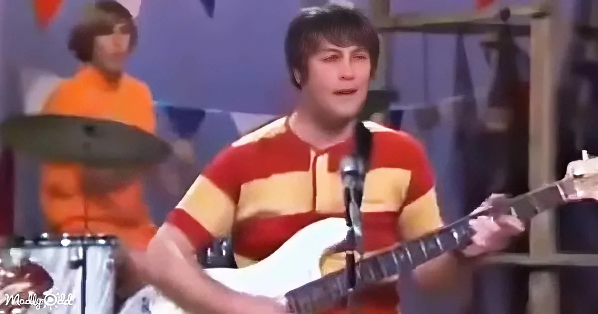 A man in a red and yellow striped shirt plays guitar on stage, singing into a microphone.