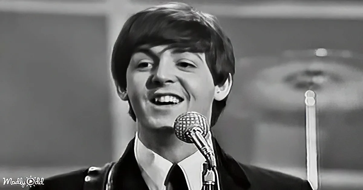 A smiling man in a suit and tie stands behind a microphone, likely performing or speaking.
