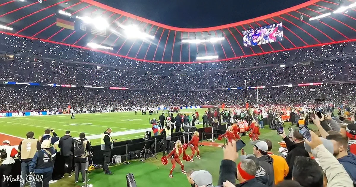 Cheerleaders in red uniforms perform on a football field surrounded by cheering fans in a brightly lit stadium.