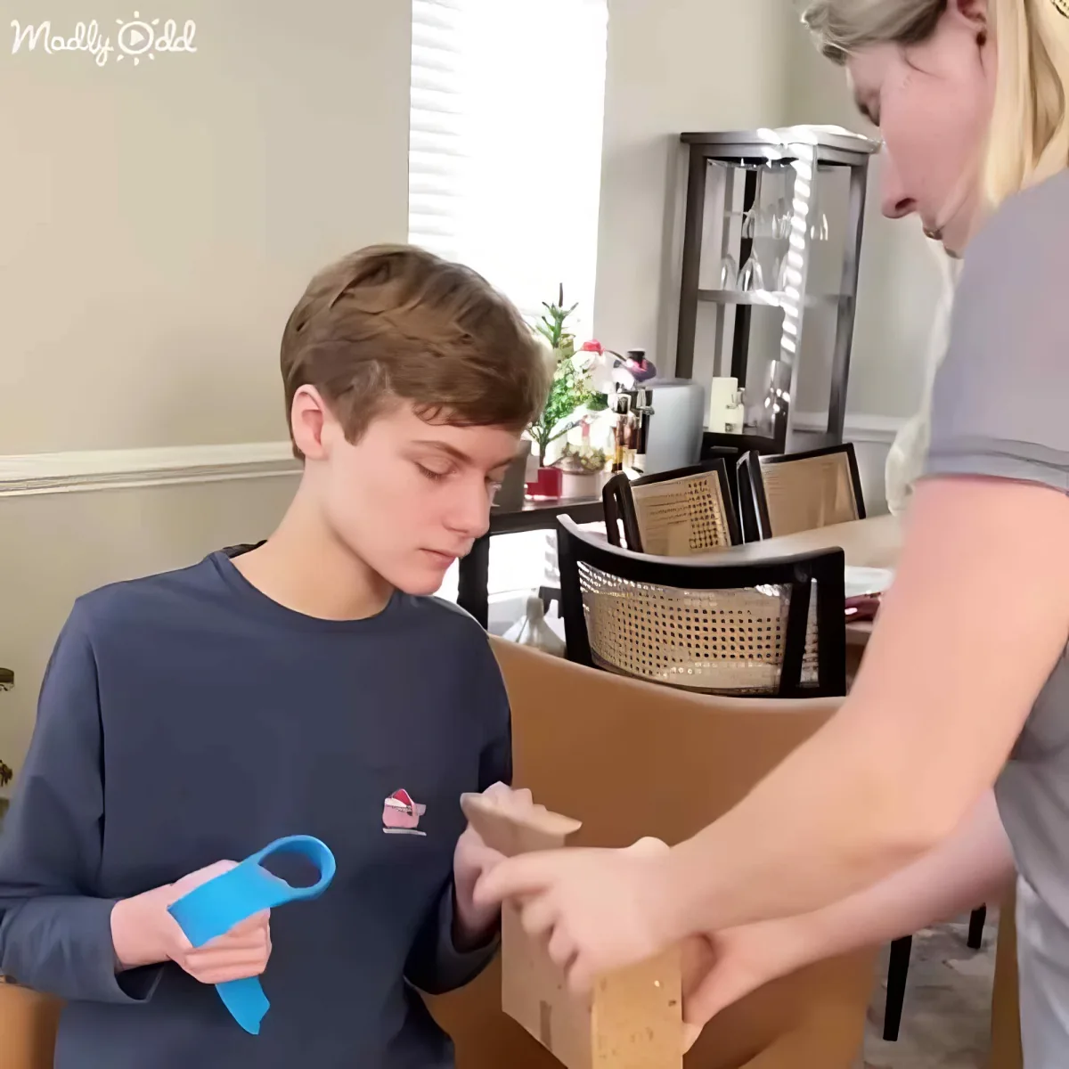A boy in a blue shirt holds a blue object while a woman in a gray shirt points at something in a brown box.