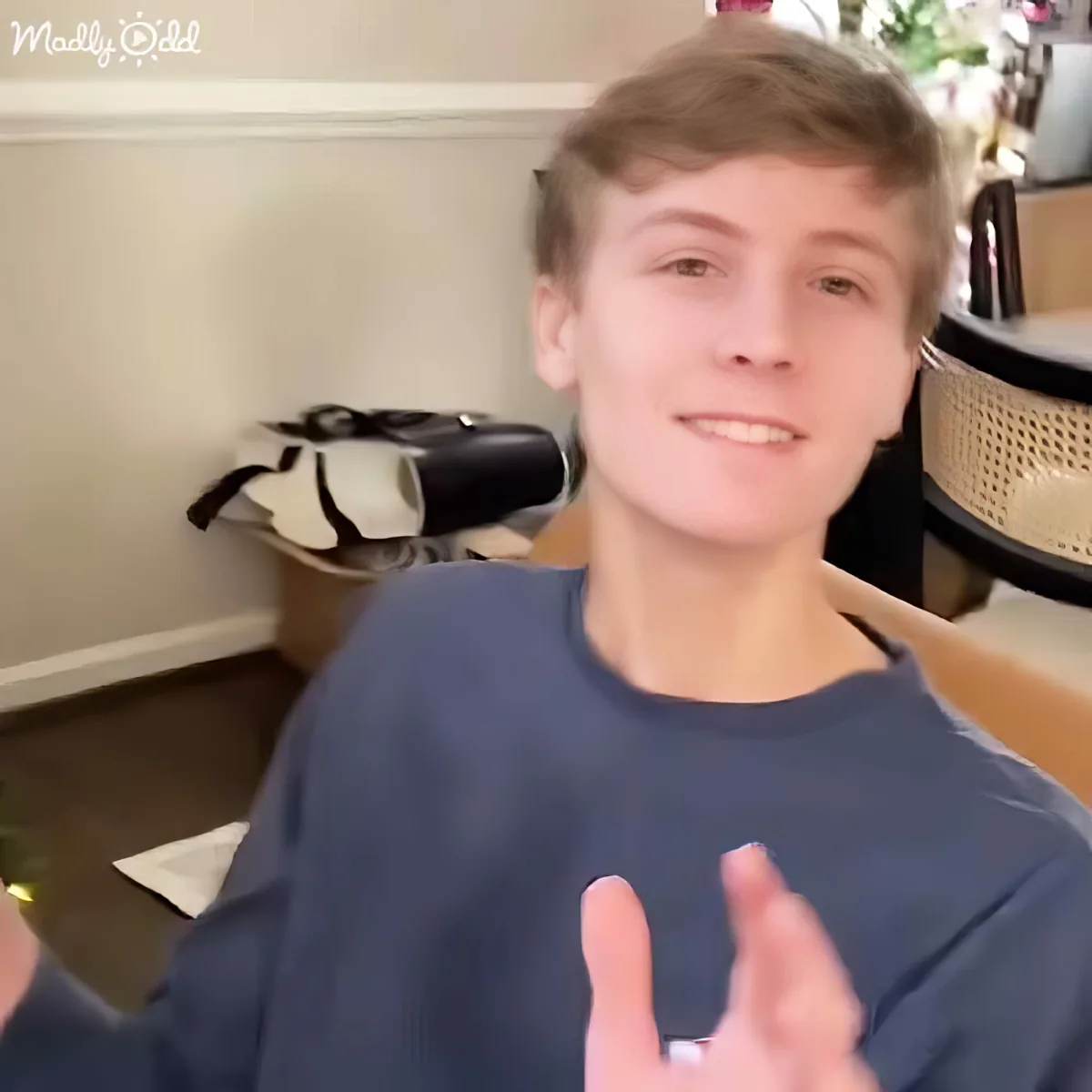 A person in a navy blue shirt is making a hand gesture, smiling, with a background showing a shoe rack and some decor.
