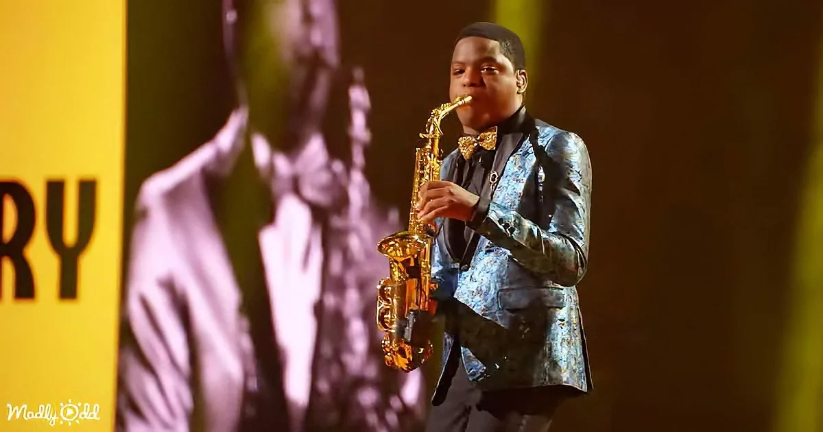 A man in a floral suit plays a saxophone on stage, with a large screen behind him.