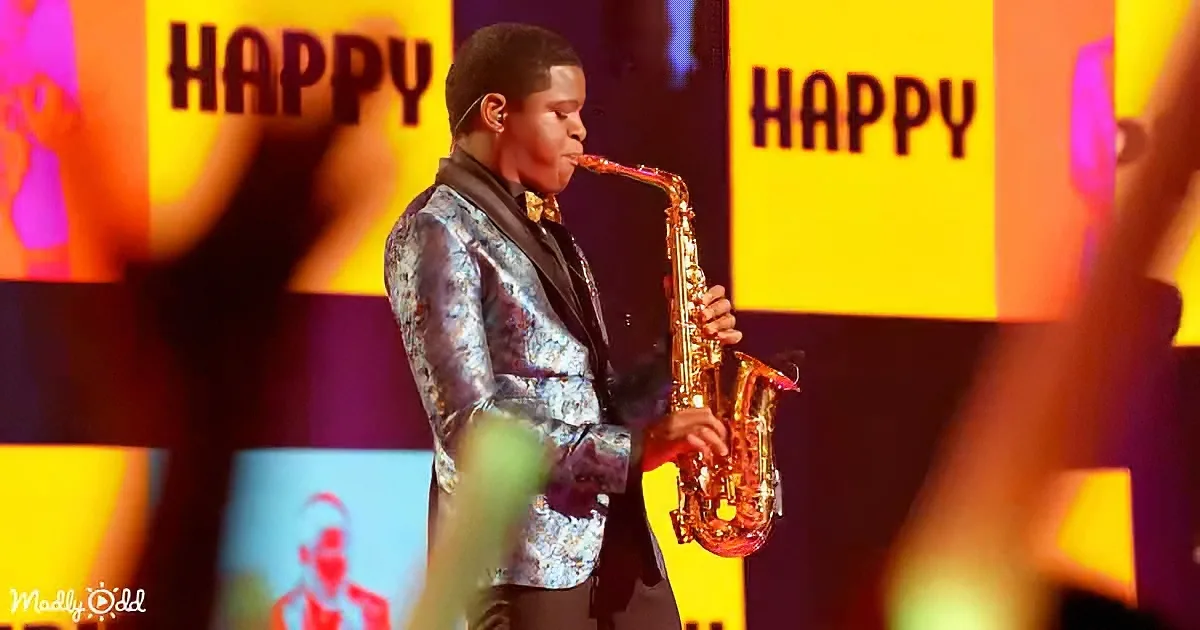 A man in a shiny jacket plays a saxophone on stage with "HAPPY" displayed in the background.