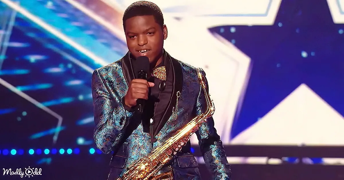 A young man in a shiny blue suit holds a saxophone and microphone on stage.