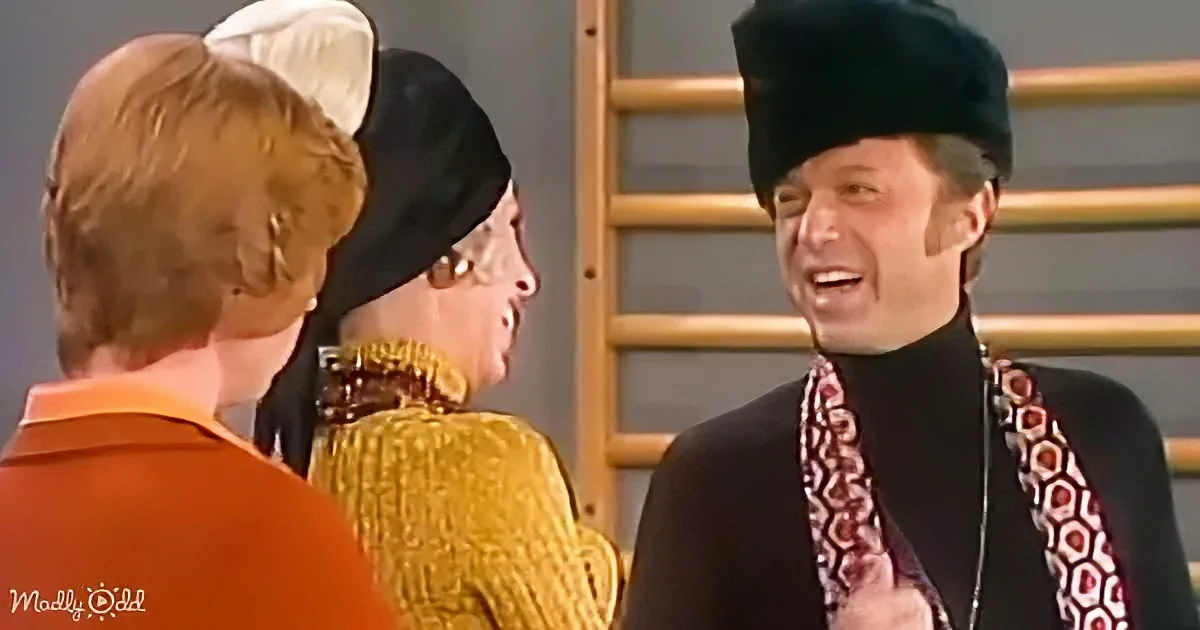 A man in a fur hat and patterned scarf is speaking to two women in a gym setting.