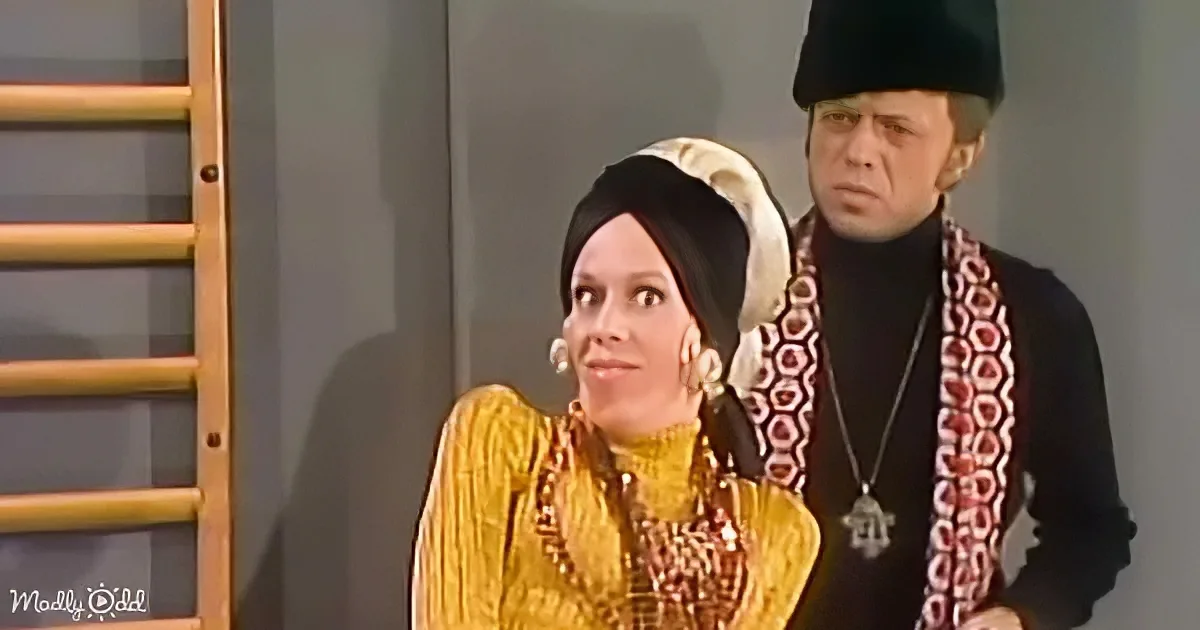 A woman in a yellow dress and headscarf looks surprised, while a man in a black outfit with a red patterned scarf stands...