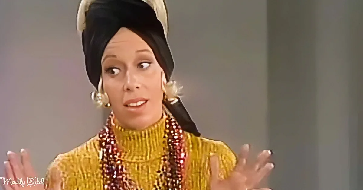 A woman in a yellow turtleneck and black headscarf gestures with her hands, wearing large earrings and beaded necklaces.