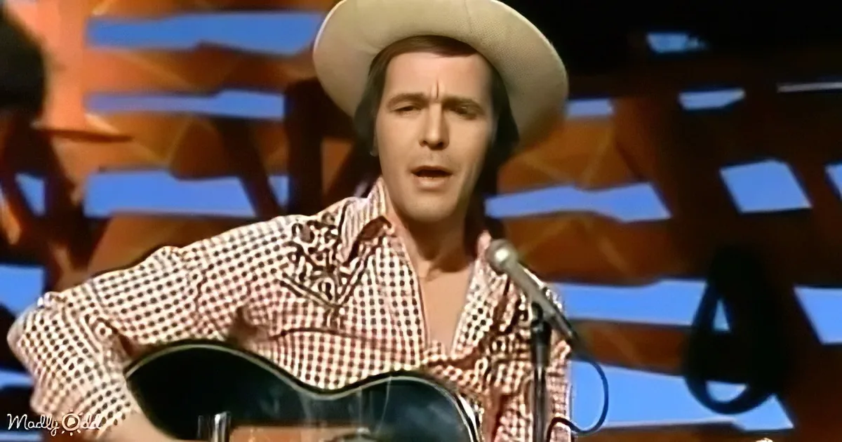 A man in a cowboy hat and checkered shirt plays guitar on stage, singing into a microphone.