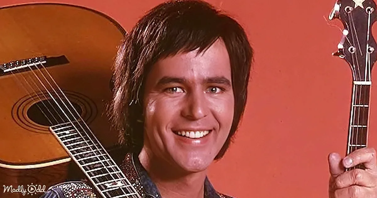A smiling man with long hair holds a guitar, with another guitar in the background against an orange wall.