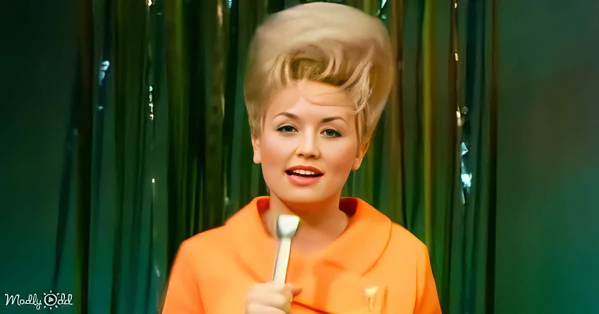 A person in an orange outfit holds a spoon, with a green curtain backdrop.