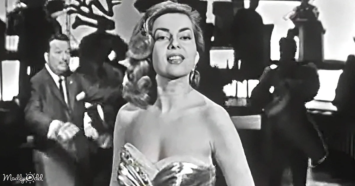 A woman in a strapless dress holds a fan, standing in a room with a man in a suit and others in the background.