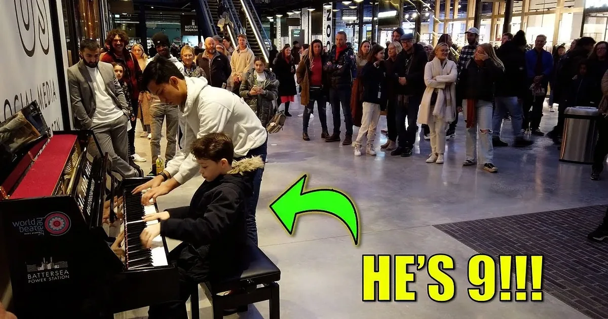 A man and boy play piano together, surrounded by a crowd in a public space.