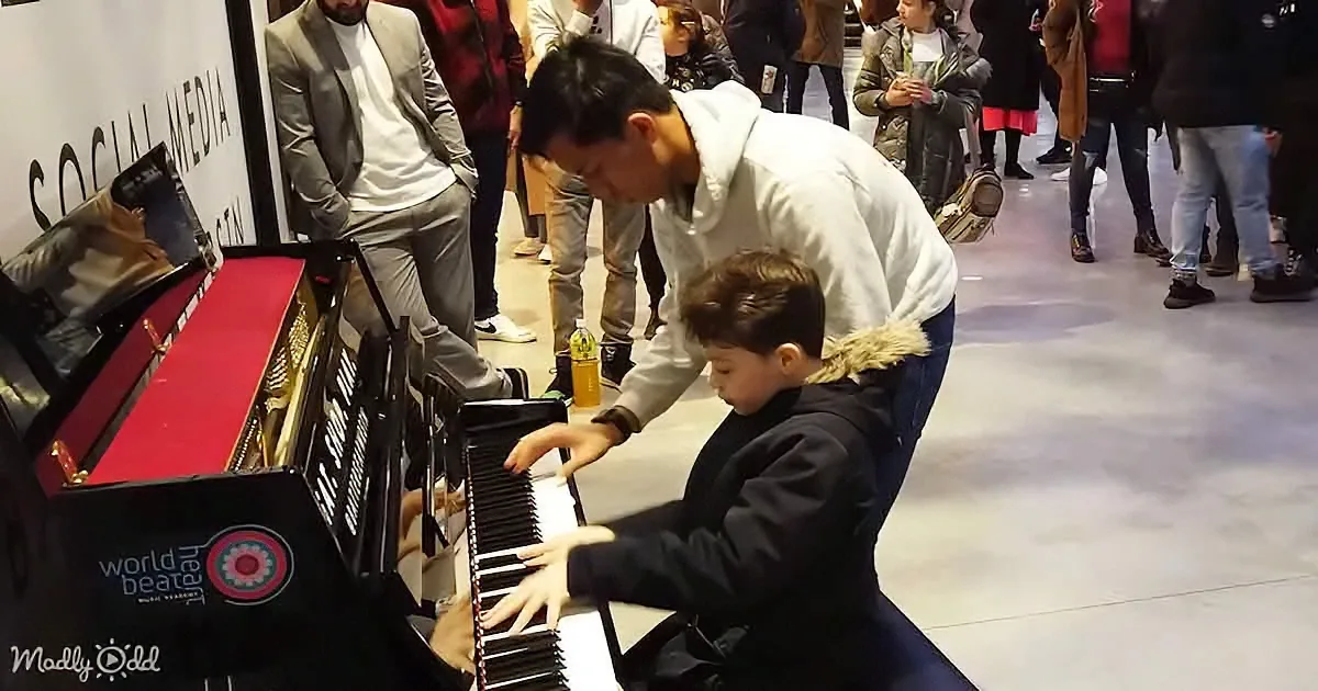 A young boy plays piano with guidance from an adult, surrounded by onlookers at a public event.
