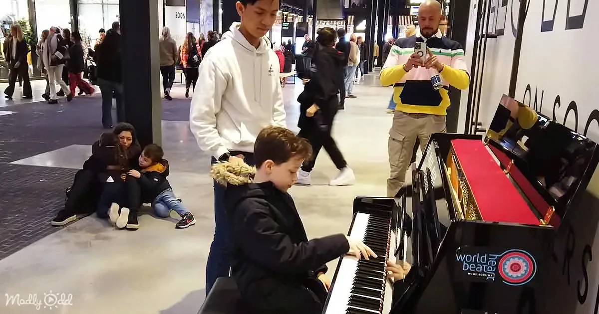 A young boy plays a piano in a busy shopping mall, surrounded by onlookers.