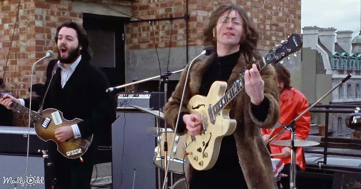 A man in a fur coat plays guitar on a rooftop, with another man singing and playing bass nearby.