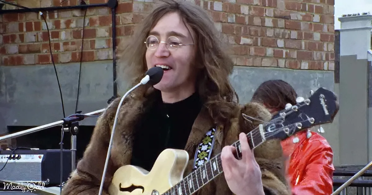 A person with long hair and glasses plays a guitar and sings into a microphone on a rooftop.
