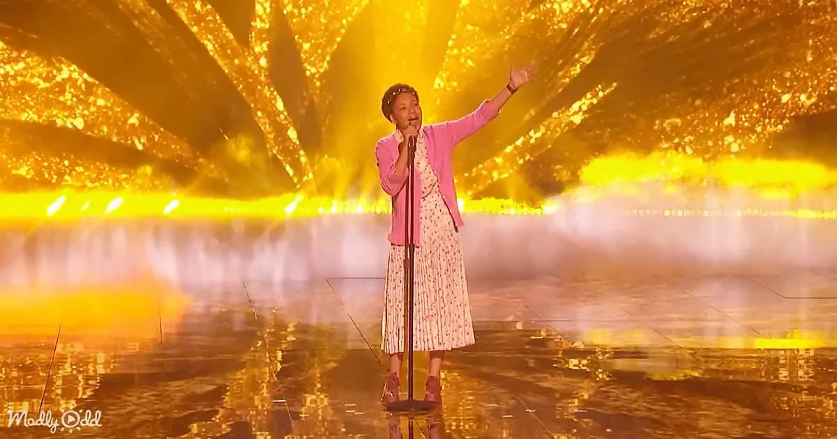 A person in a pink outfit stands on a stage with a microphone, pointing upwards, surrounded by golden lights and mist.