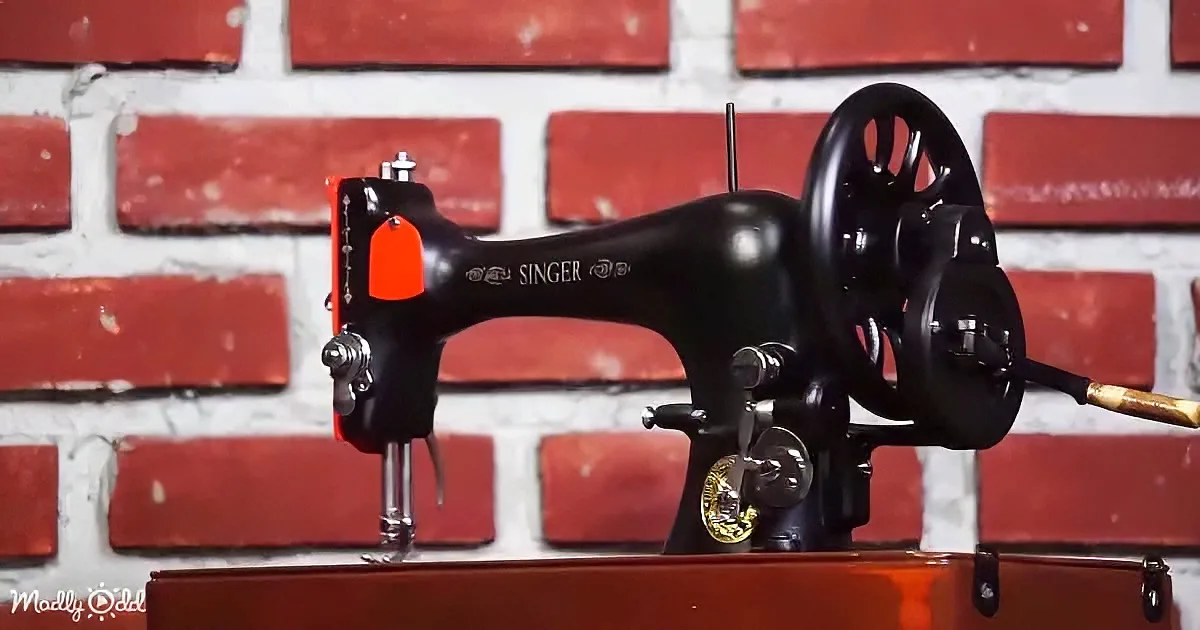 A vintage Singer sewing machine with a red tag and black body sits on a wooden surface against a red brick wall.