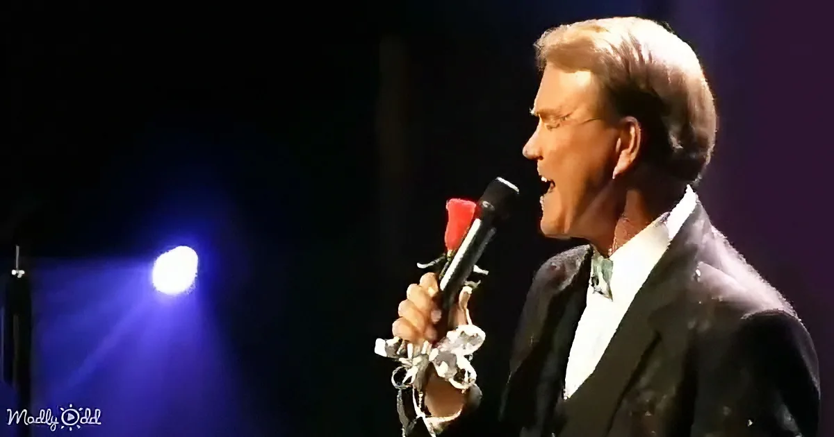 A man in a suit sings passionately into a microphone on stage, illuminated by blue light.