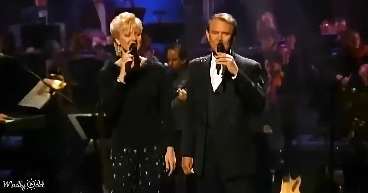 A man and woman in black attire sing on stage with microphones, surrounded by an orchestra.