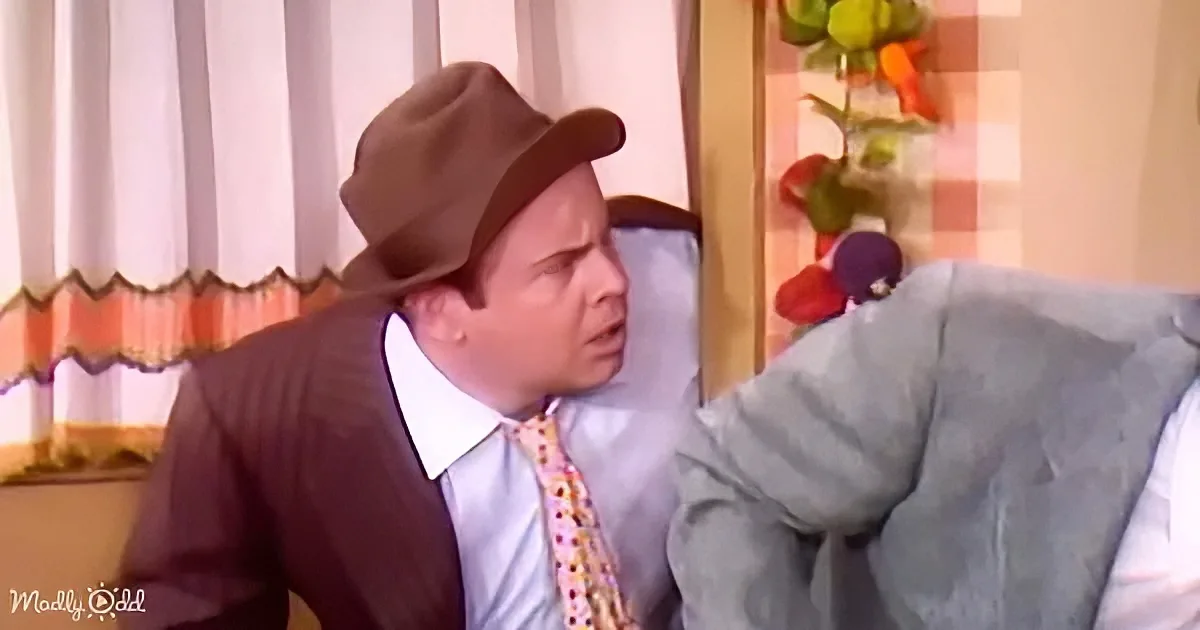 A man in a brown hat and suit is sitting on a couch, looking surprised.