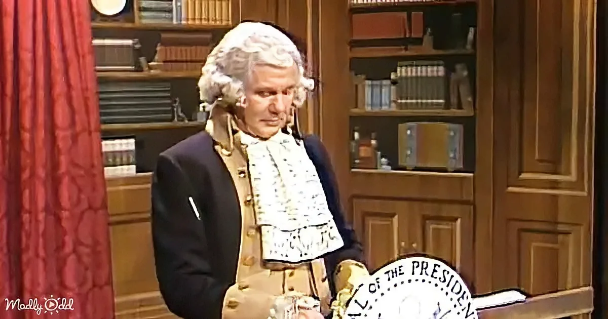 A man in colonial attire stands in front of a bookshelf, holding a presidential seal.