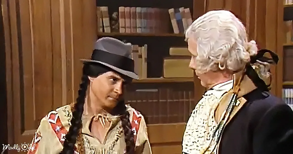 A man in a Native American outfit and a man in a colonial costume are having a conversation in a room with bookshelves.