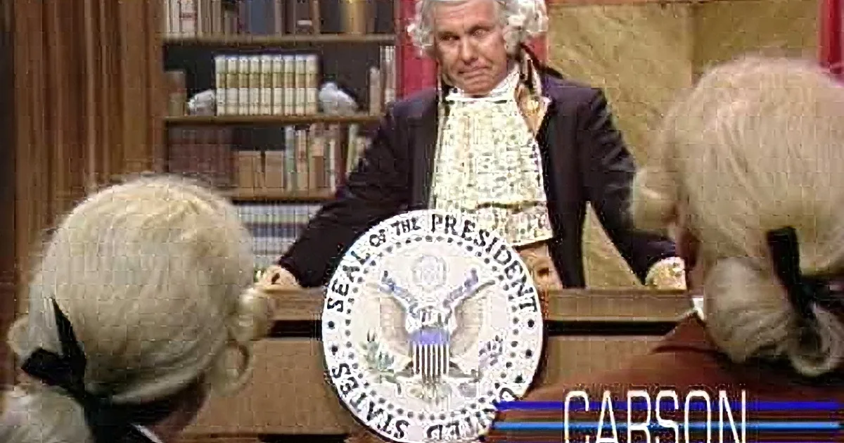 A person in colonial attire sits at a desk with the presidential seal, smiling.
