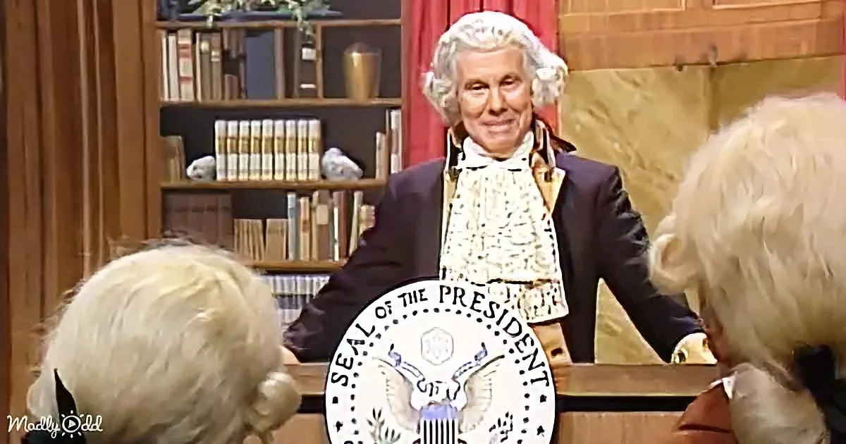A person dressed as George Washington stands behind a desk with the Presidential Seal, addressing two seated individuals.