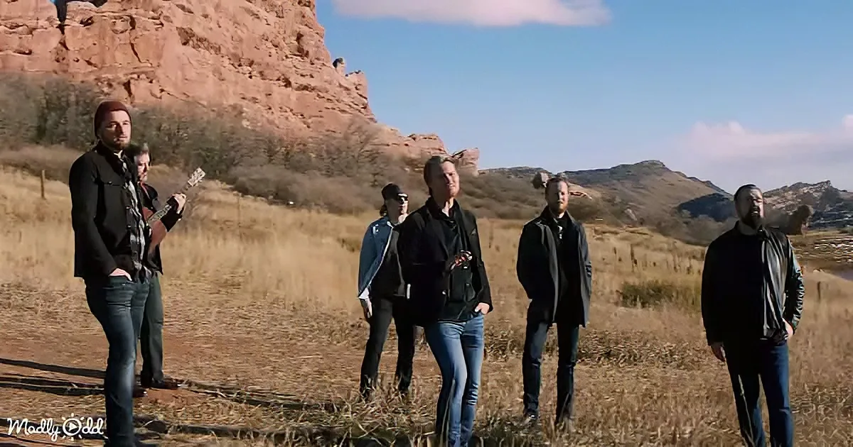 A group of six people stands in a field with a red rock formation in the background.