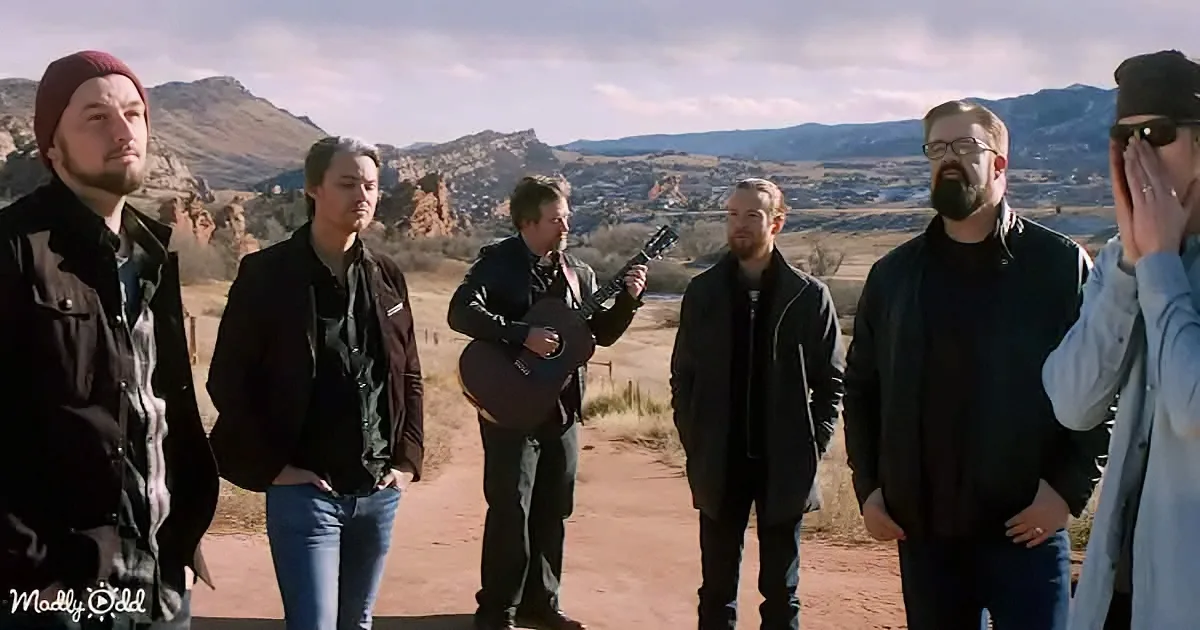 A man plays guitar while five others stand in a desert landscape, watching.