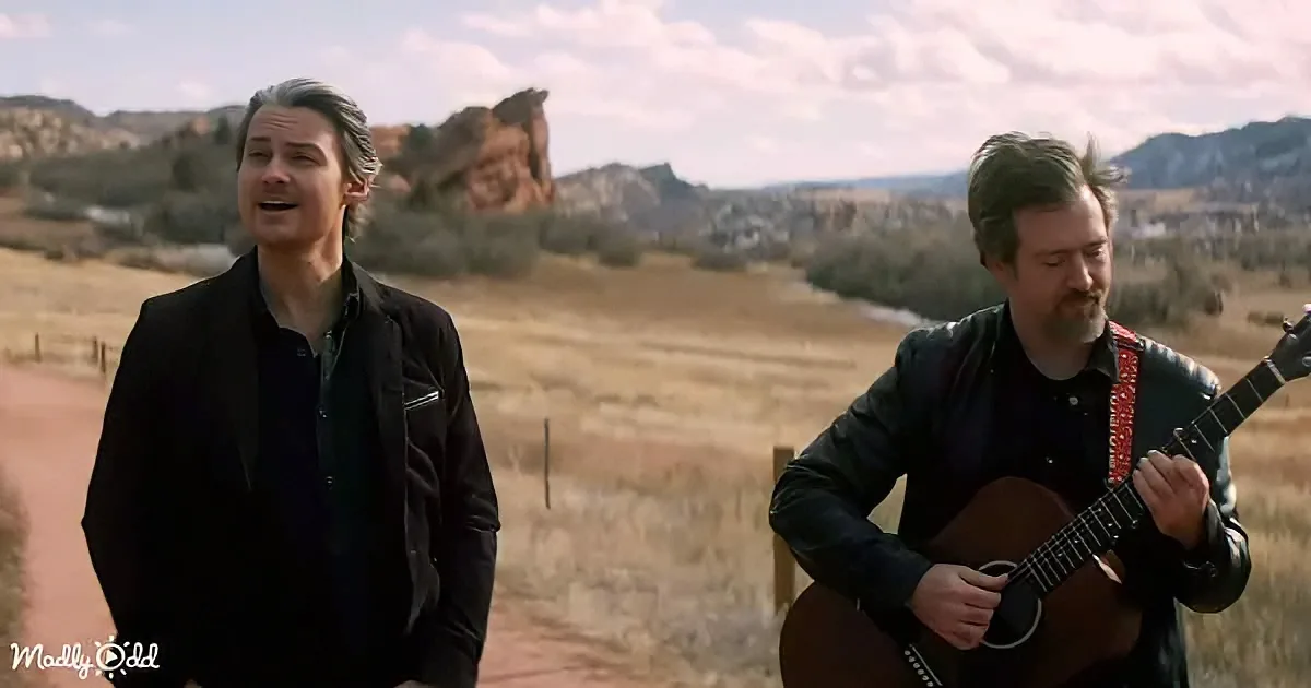 A man sings while another plays guitar in a scenic, open landscape with mountains in the background.
