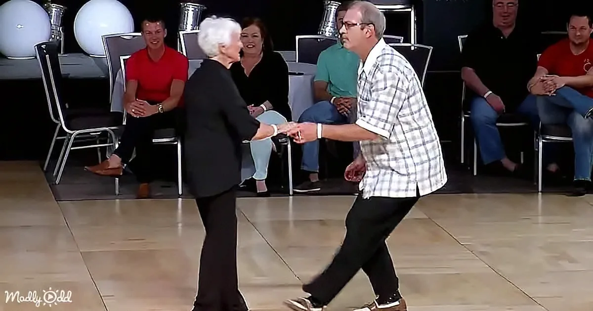 An elderly couple dances on a wooden floor, surrounded by seated spectators in a dimly lit room.