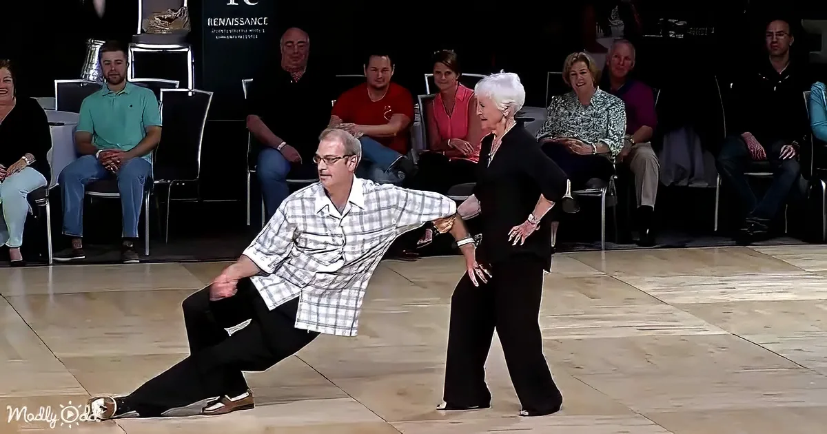 An older couple dances gracefully on a wooden floor, surrounded by an audience in a dimly lit room.