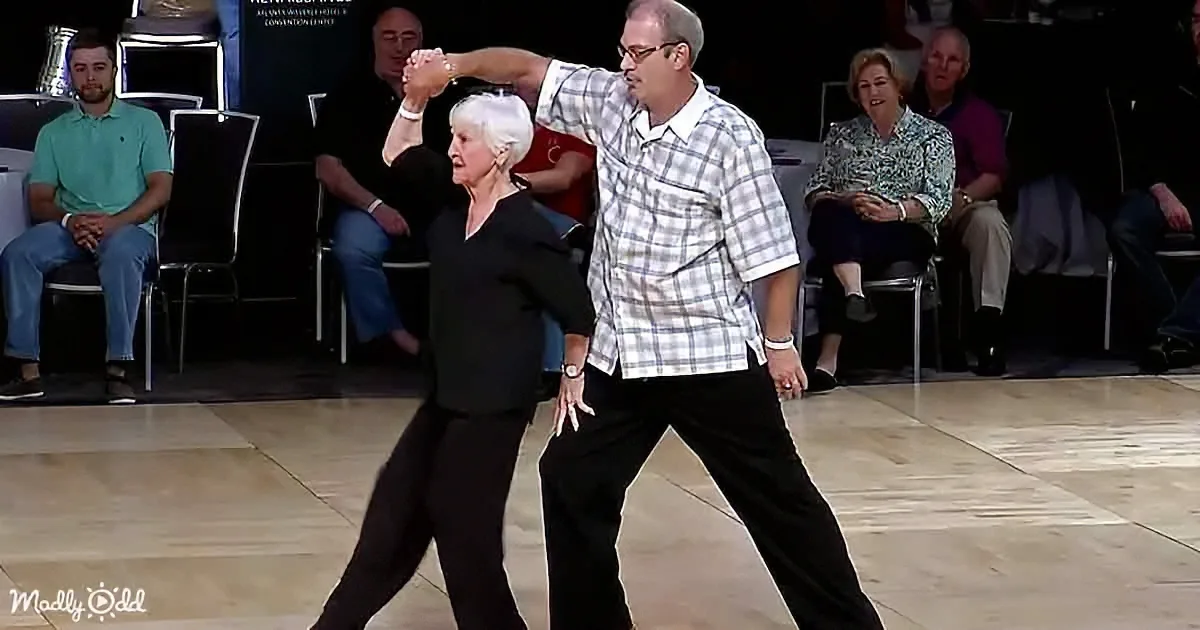 An older couple dances gracefully on a stage, holding hands, with an audience watching in the background.