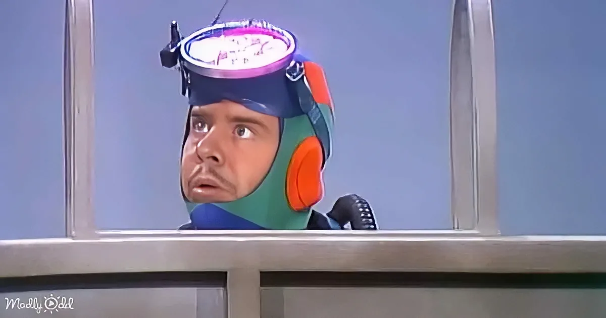 A man in a colorful headpiece with a watch on it looks surprised, behind a metal railing.